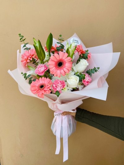 A bright bouquet with coral gerberas, white roses, pink carnations, and unopened lily buds wrapped in soft pink paper.