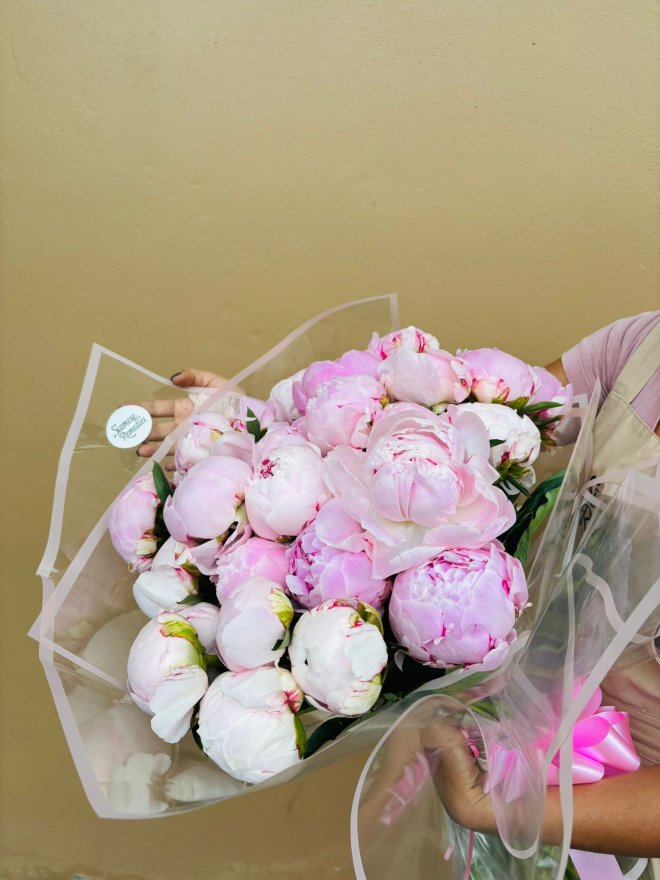 A large bouquet of soft pink peonies wrapped in translucent paper and tied with a pink ribbon.