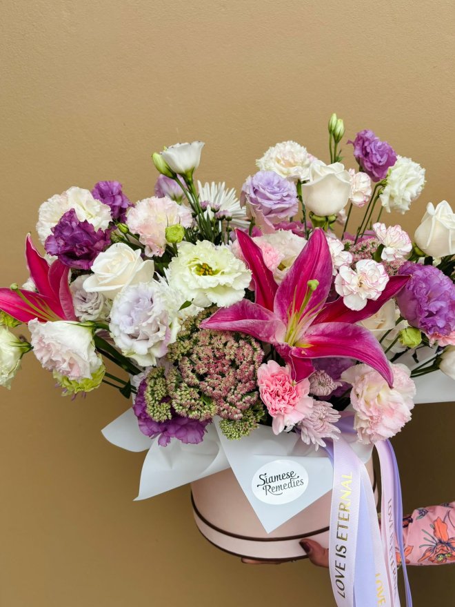 A pastel hat box arrangement with pink lilies, lisianthuses, roses, and carnations in soft pink, purple, and white tones.