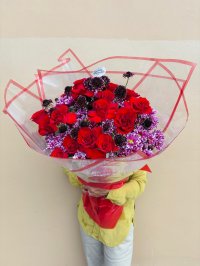 A bouquet of red roses, burgundy scabiosa, and purple chrysanthemums wrapped in clear red-trimmed paper.