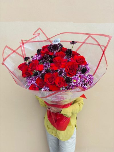 A bouquet of red roses, burgundy scabiosa, and purple chrysanthemums wrapped in clear red-trimmed paper.