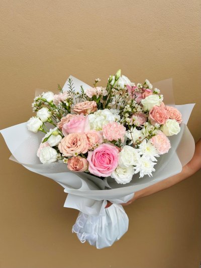 A pastel bouquet of peach, pink, and white roses, carnations, lisianthus, chrysanthemums, and wax flowers wrapped in frosted white paper.