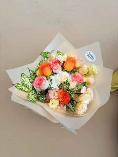 A cheerful bouquet of orange and peach roses with carnations, chrysanthemums, lisianthuses, and greenery, wrapped in soft beige paper.