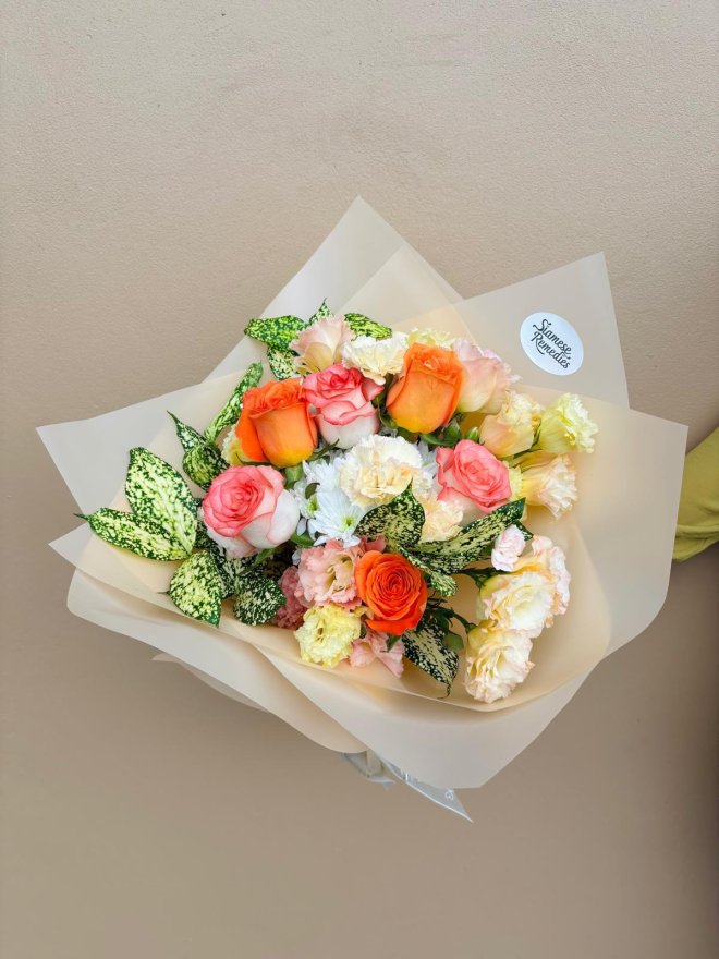 A cheerful bouquet of orange and peach roses with carnations, chrysanthemums, lisianthuses, and greenery, wrapped in soft beige paper.