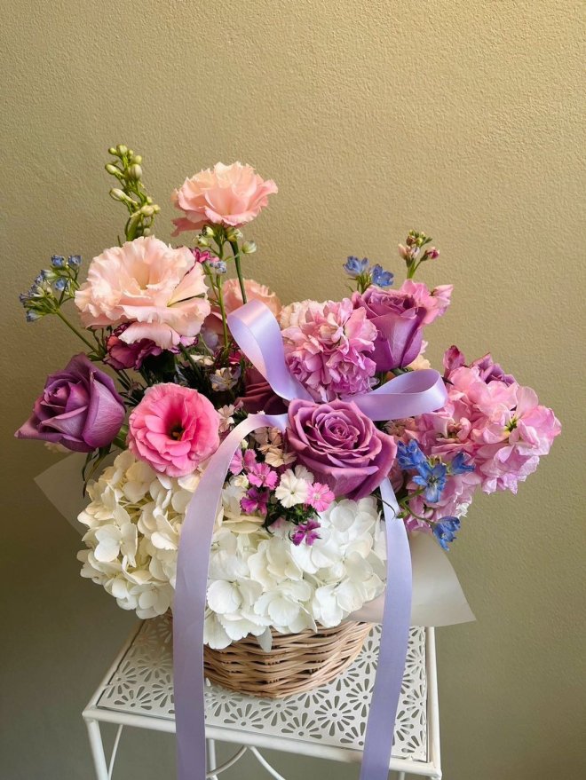 Pastel flower basket with lavender roses, blush lisianthus, white hydrangeas, blue and pink accent flowers, tied with lavender ribbon.