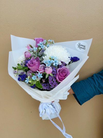 A white-wrapped bouquet with purple roses, white chrysanthemums, blue forget-me-nots, and violet statice flowers.