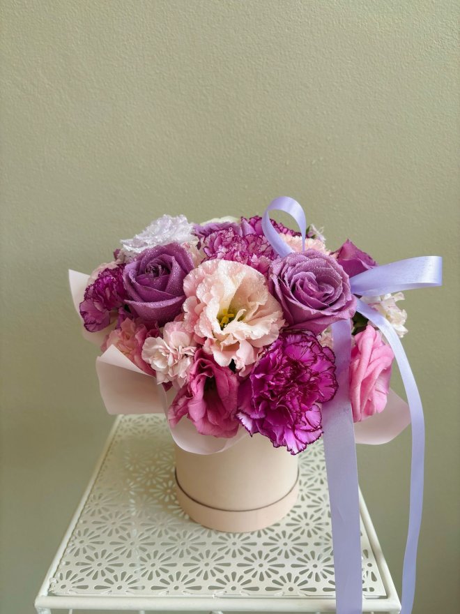 Box arrangement with purple roses, pink carnations, lisianthus, and peonies tied with a lavender ribbon.