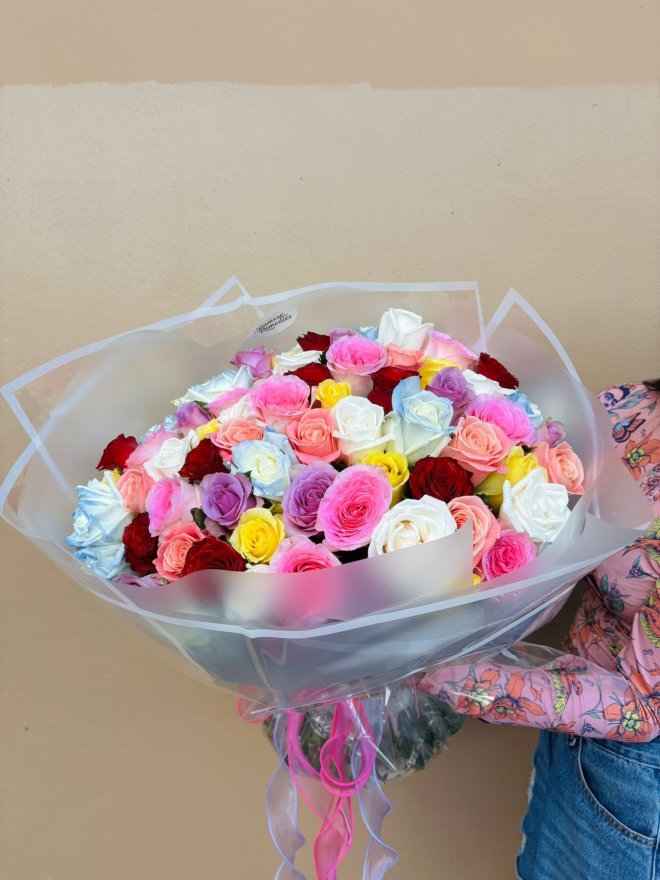 A large bouquet of multicolored roses in red, pink, yellow, white, peach, purple, and blue, wrapped in sheer white paper.