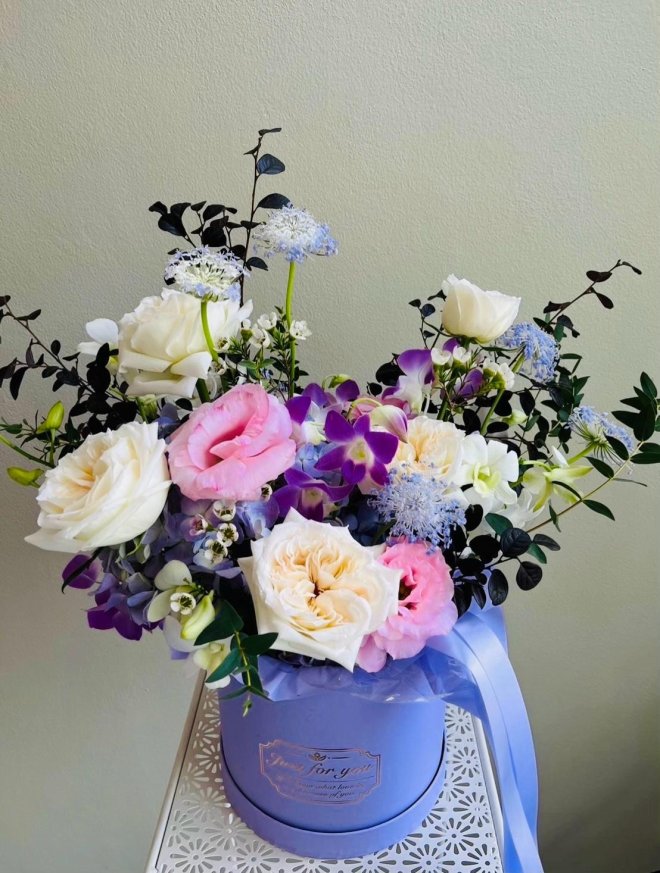 Hatbox bouquet of white roses, pink lisianthus, purple orchids, blue lace flowers, and hydrangeas with light blue ribbon
