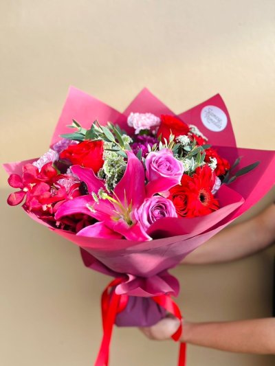 A bouquet of red roses, pink lilies, orchids, and carnations wrapped in deep pink paper with a red ribbon.