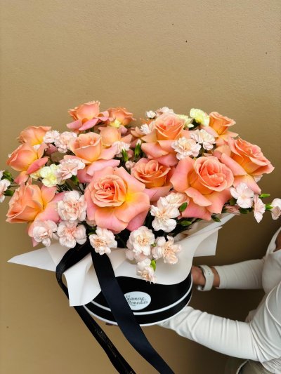 A black hat box filled with peach roses and blush carnations, tied with black and white ribbons.