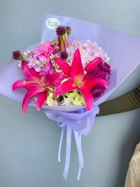 A vibrant hand-tied bouquet with pink lilies, carnations, hydrangeas, and purple alliums wrapped in lilac paper.