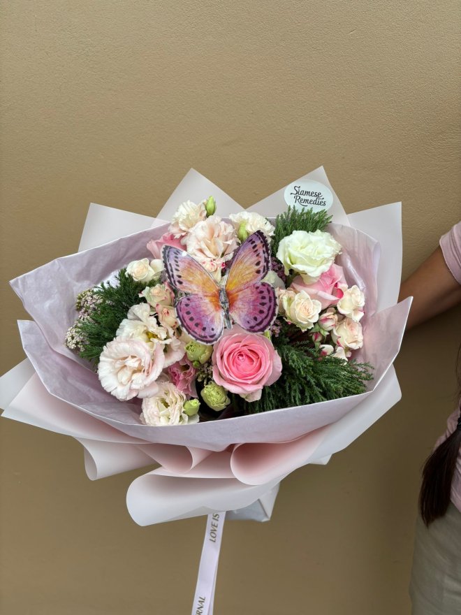 A pastel bouquet featuring pink roses, lisianthuses, and carnations with a decorative butterfly on top.