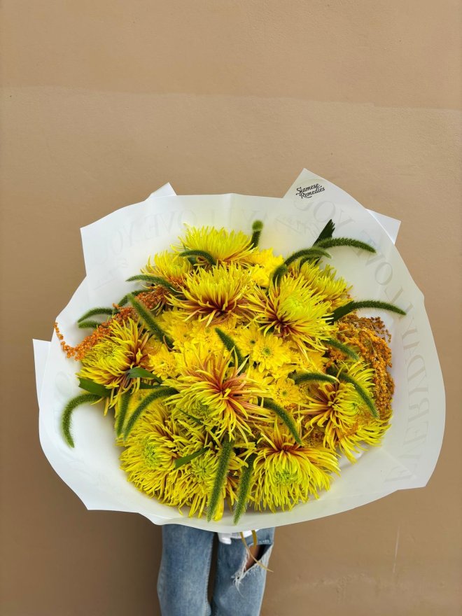 A bouquet of yellow spider chrysanthemums with golden fillers and greenery, wrapped in white paper.