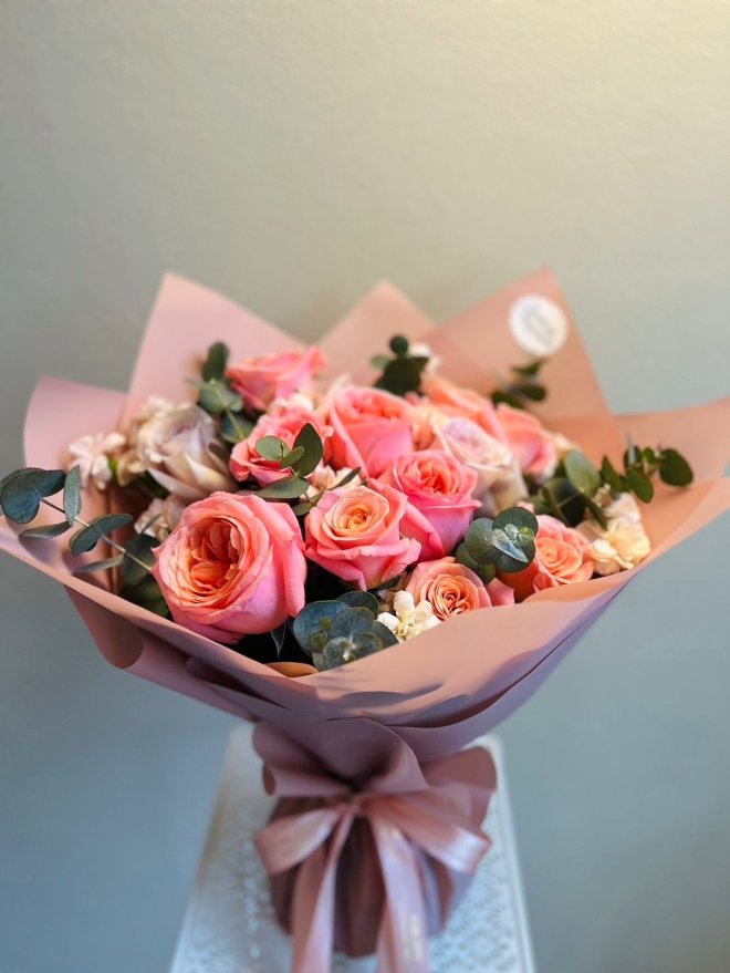 A bouquet of coral, peach, and blush roses with greenery, wrapped in rose-pink paper.