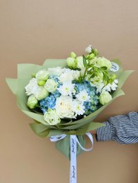 A fresh bouquet with white roses, lisianthus, blue hydrangeas, and chrysanthemums wrapped in green paper.