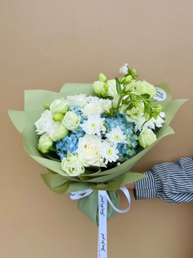 A fresh bouquet with white roses, lisianthus, blue hydrangeas, and chrysanthemums wrapped in green paper.