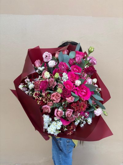 A dramatic bouquet of crimson, pink, and blush roses, lisianthuses, and chrysanthemums wrapped in deep red paper.