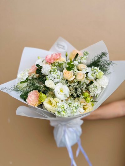 A soft bouquet of white, cream, and peach roses, lisianthuses, carnations, and dry flowers wrapped in white paper.
