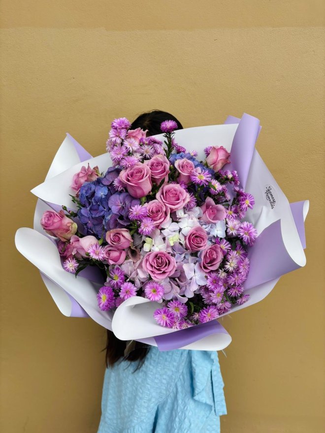 A bouquet of purple roses, blue hydrangeas, and lilac asters wrapped in white and lavender paper.