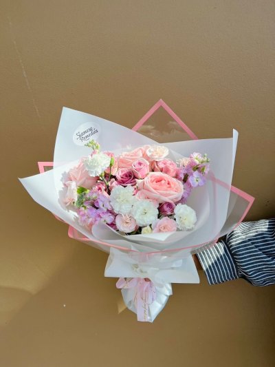 A pastel bouquet featuring pink roses, carnations, lisianthus, and stock flowers, wrapped in white with a pink edge.
