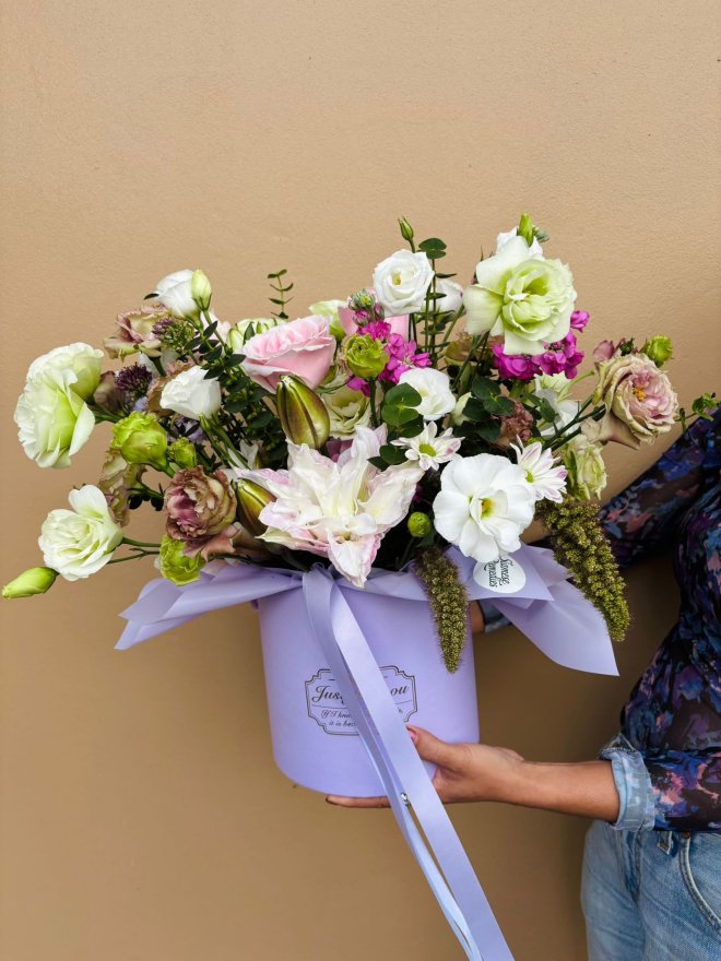 A lavender hat box with white, pink, green, and lilac flowers including lilies, lisianthuses, and roses.
