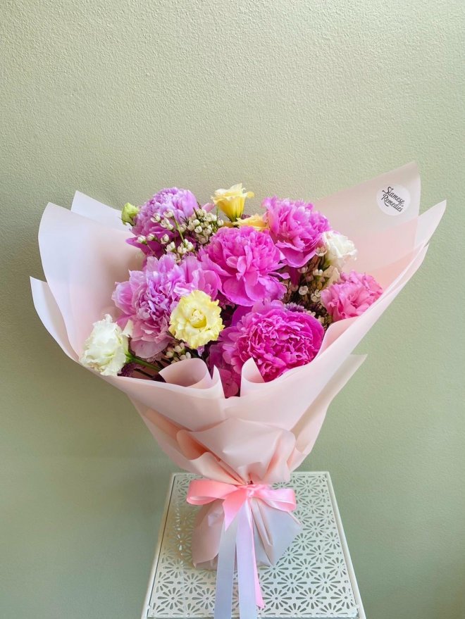 Bouquet of pink and lilac peonies, yellow lisianthus, baby’s breath, and white blooms wrapped in blush paper with pink and lavender ribbons
