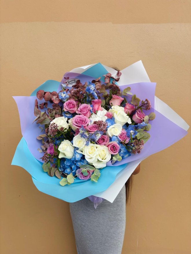 A bouquet of pastel roses and blue hydrangeas with rustic foliage, wrapped in lilac and sky-blue paper.