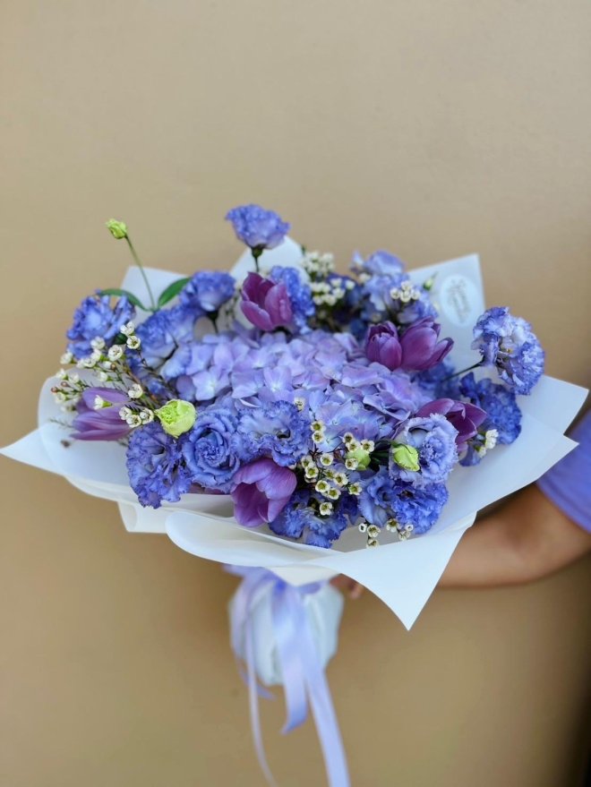 A lavender bouquet with hydrangeas, lisianthus, carnations, tulips, and wax flowers in soft purple and blue tones.