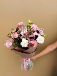 A bouquet of pink lisianthus, stock, white carnations, and deep burgundy blooms wrapped in soft pink and brown paper.