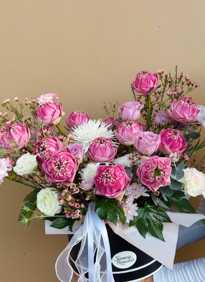 A floral arrangement in a box featuring pink folded lotus flowers, white lisianthus, roses, chrysanthemums, and wax flowers.