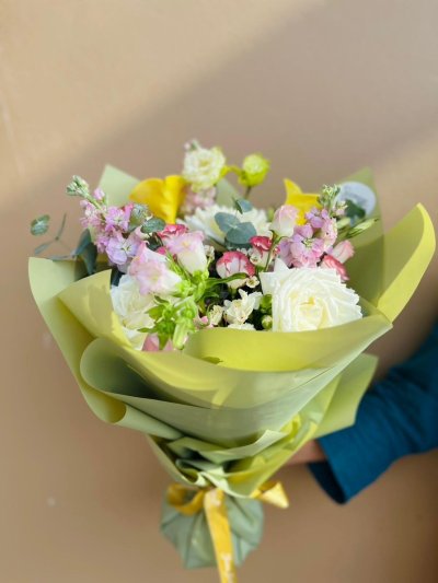 A vibrant bouquet of yellow calla lilies, white roses, pink lisianthus, and mixed blooms wrapped in olive green paper with a yellow ribbon.