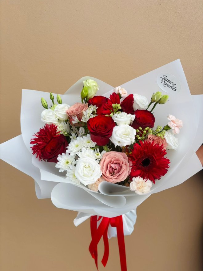 A white-wrapped bouquet featuring red gerberas, roses, white lisianthus, chrysanthemums, and blush carnations.