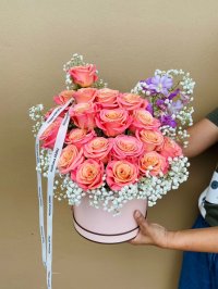 A coral and peach rose arrangement with baby's breath and lilac blooms in a pink hatbox.