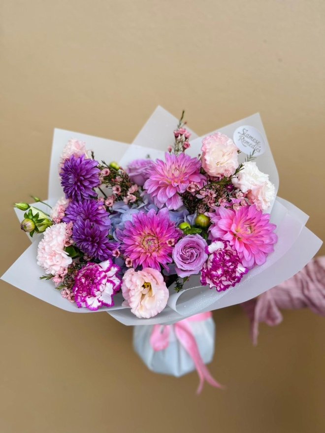 A wrapped bouquet featuring purple and pink dahlias, carnations, lisianthuses, roses, and wax flowers.