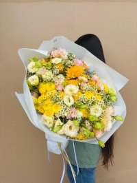 A large bouquet featuring yellow chrysanthemums, lisianthus, peach carnations, daisies, and a sunflower wrapped in white paper.