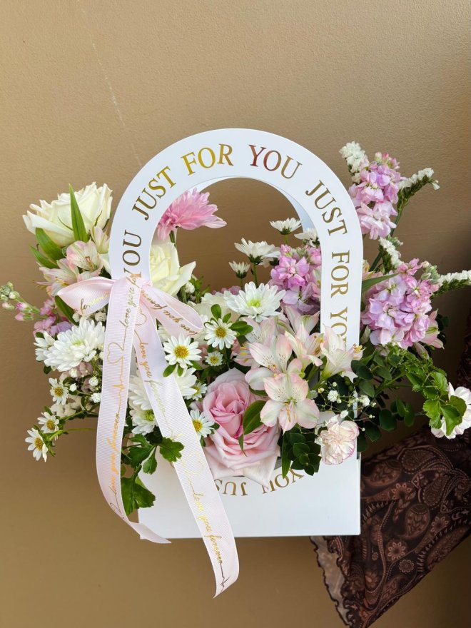 A white carry bag filled with pastel flowers including pink roses, white daisies, stock, and alstroemeria, tied with soft pink ribbon.