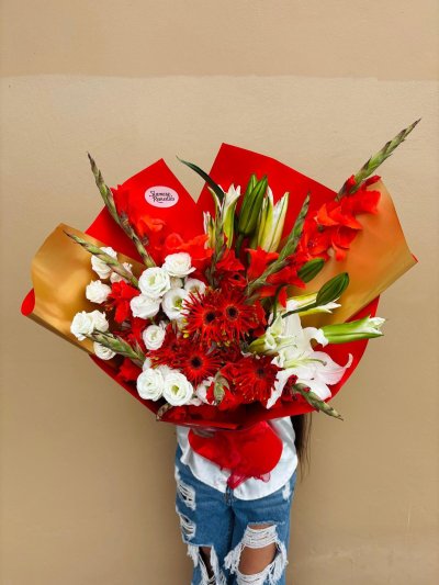 A vibrant bouquet of red gerberas, white lilies, lisianthuses, and gladiolus wrapped in red and gold.