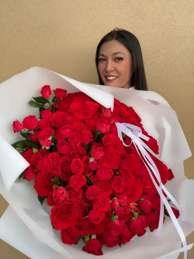 A large bouquet of red roses wrapped in white paper with white ribbon, held by a smiling woman.
