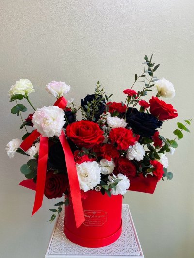 Red, white, and black floral arrangement with roses, carnations, and lisianthus in a red hatbox with red satin ribbon