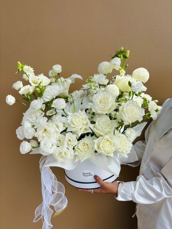 A white floral arrangement with roses, lisianthus, chrysanthemums, and lilies in a round box with sheer white ribbon.