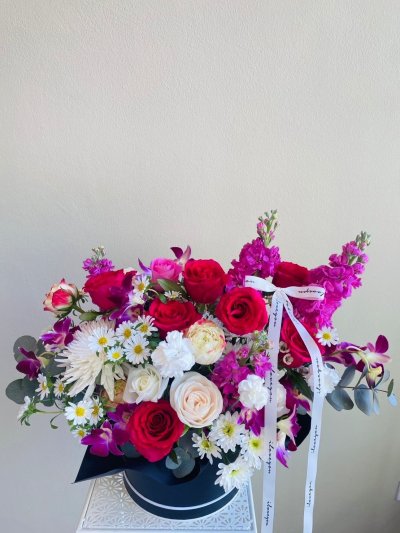 A vibrant floral hatbox arrangement with red roses, purple orchids, carnations, chrysanthemums, and daisies in a black box with white ribbon.
