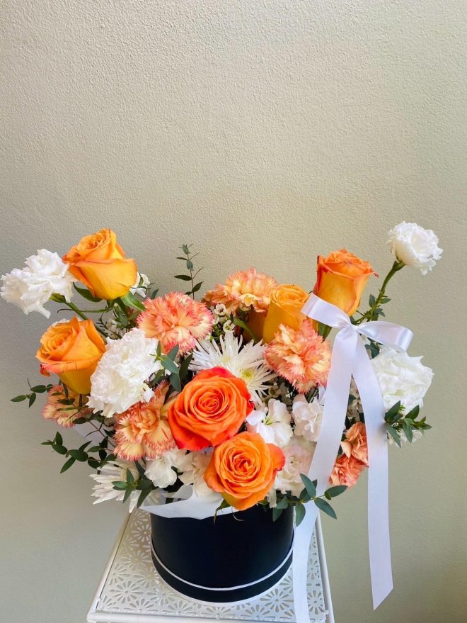 Floral arrangement of orange roses, white carnations, mums, and peachy carnations in a black hatbox with white satin ribbon