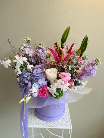 A pastel floral box featuring lilies, roses, orchids, carnations, and hydrangeas in pink, purple, and white.