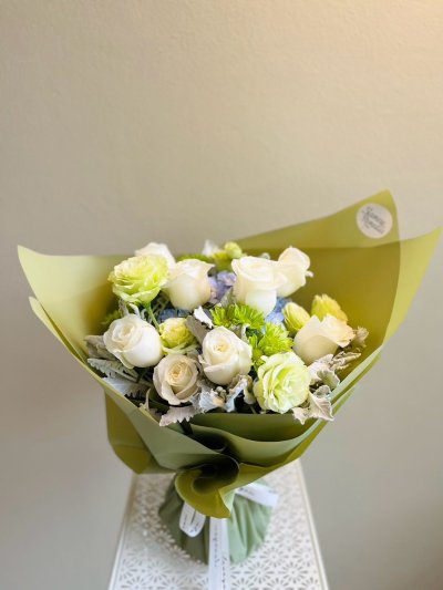 Bouquet of white roses, green carnations, and dusty miller foliage wrapped in olive green paper with a white ribbon