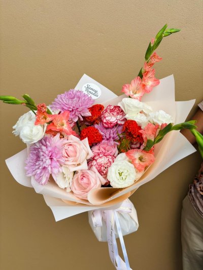 A vibrant bouquet featuring pink roses, gladiolus, red and bi-color carnations, lisianthus, and purple chrysanthemums wrapped in peachy-white paper.