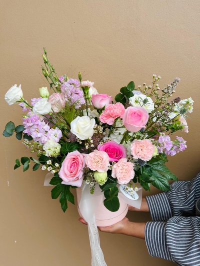 A pastel hatbox arrangement featuring pink roses, carnations, lisianthuses, and wax flowers.