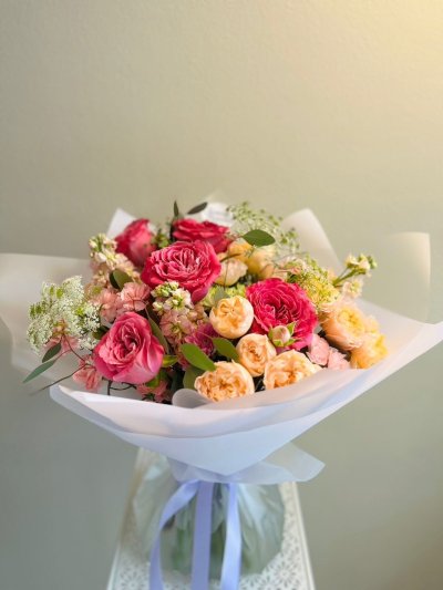 A bouquet of raspberry pink and peach roses with greenery, wrapped in soft white paper and tied with a lilac ribbon.