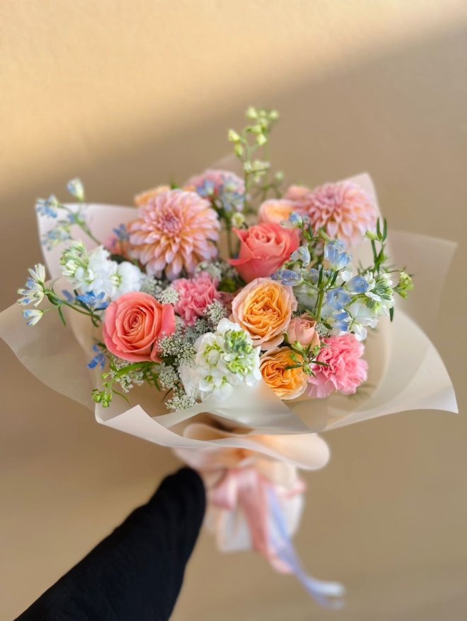 A pastel bouquet of dahlias, roses, carnations, and dry flowers in peach, pink, blue, and white, wrapped in sheer paper with ribbon.
