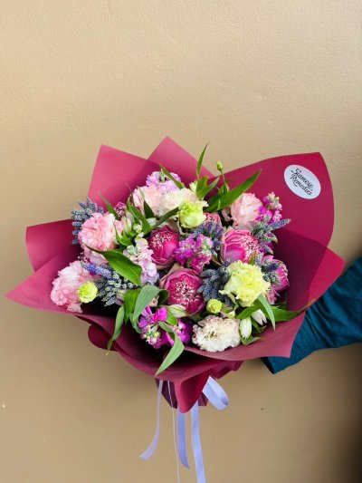 A vibrant bouquet with pink lotus, pink carnations, white lisianthus, lavender stock, and veronica flowers wrapped in berry-red paper.
